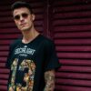 A stylish young man with tattoos wearing sunglasses and a graphic t-shirt poses confidently.