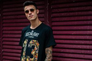 A stylish young man with tattoos wearing sunglasses and a graphic t-shirt poses confidently.