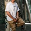 Stylish man posing against graffiti column in urban setting, wearing white t-shirt and cargo pants.