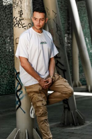 Stylish man posing against graffiti column in urban setting, wearing white t-shirt and cargo pants.