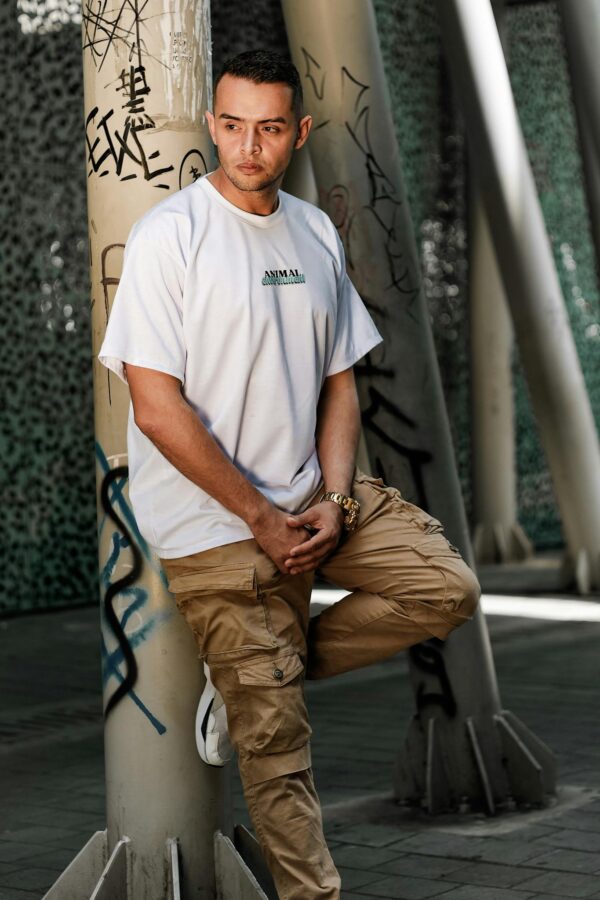 Stylish man posing against graffiti column in urban setting, wearing white t-shirt and cargo pants.