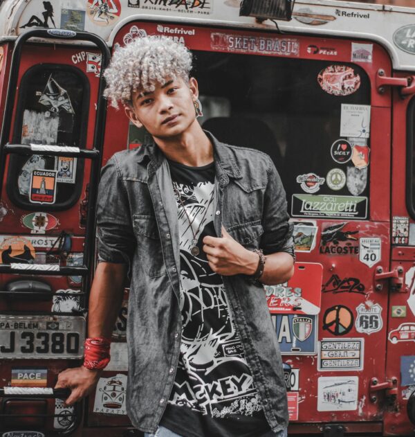 Stylish young man with curly hair poses against a sticker-covered van showcasing urban street fashion.